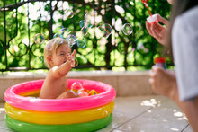 Wet Baby Sits In A Small Inflatable Pool And Catches Soap Bubbles That Mom Lets Out