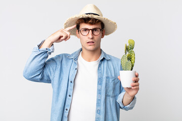 Wall Mural - young handsome man feeling confused and puzzled, showing you are insane. farmer holding a decorative cactus