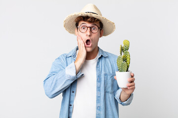 Wall Mural - young handsome man feeling shocked and scared. farmer holding a decorative cactus