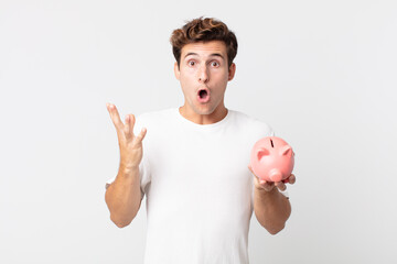 Wall Mural - young handsome man feeling extremely shocked and surprised and holding a piggy bank