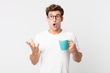 Wall Mural - young handsome man feeling extremely shocked and surprised and holding a coffee cup