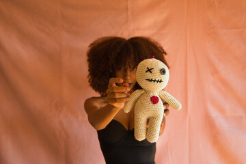 Young Latina woman in black dress holding voodoo doll in foreground on pink background.