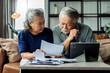 old retired asian senior couple checking and calculate financial billing together on sofa involved in financial paperwork, paying taxes online using e-banking laptop at living room home background