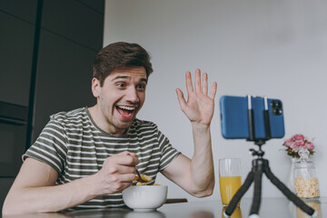 Wall Mural - Young friendly amazed excited caucasian man in striped t-shirt use mobile cell phone say hello eat breakfast muesli cereals talk by video call waving hand sit by table in light kitchen at home alone