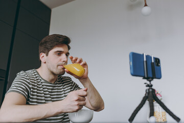 Wall Mural - Bottom view young caucasian man 20s wear striped t-shirt use mobile cell phone watching movie film eat breakfast muesli cereals drink citrus orange juice sit by table in light kitchen at home alone.