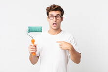 Young Handsome Man Looking Shocked And Surprised With Mouth Wide Open, Pointing To Self And Holding A Roller Paint