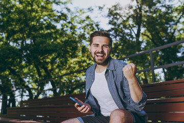 Wall Mural - Young overjoyed man in blue shirt shorts sit on bench use mobile phone do winner gesture clench fist rest relax in spring green city sunshine park outdoor on nature Urban leisure lifestyle concept.