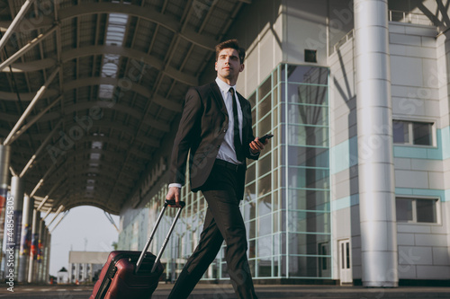 Bottom side view traveler businessman young man in black suit walk go outside at international airport terminal with suitcase use mobile phone book taxi order hotel Air flight business trip concept