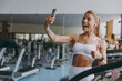 Young sporty athletic sportswoman woman in white sportswear earphones listen music do selfie shot on mobile cell phone show thumb up warm up training treadmill in gym indoors Workout sport concept