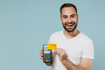 Young happy man 20s in blank print design white t-shirt hold wireless modern bank payment terminal to process acquire credit card payments isolated on plain pastel light blue color background studio