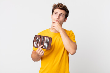 Wall Mural - young handsome man thinking, feeling doubtful and confused and holding a nails tools case