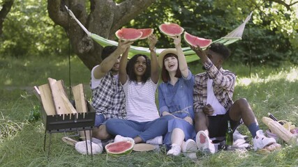 Wall Mural - Happy multi ethnic friends enjoying seasonal tasty watermelon during picnic at summer garden, raising up slices. Four young people sitting on grass and having fun on picnic