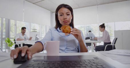 Latin businesswoman sitting at desk with coffee using laptop and eating croissant.