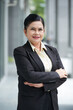 © DragonImages - Positive aged female entrepreneur crossing arms and looking at camera when standing outside the office building