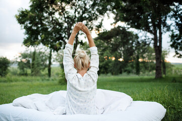 Young blonde woman wake up wear pajamas sitting in bed on white blanket outdoors over green nature background. Good morning. Back view. Relaxation. Summer vacation season.