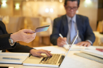 Wall Mural - Businesswoman making online order via laptop and paying with credit card when her colleague signing documents in background