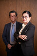 © DragonImages - Portrait of smiling senior business couple crossing arms and looking at camera when standing against wooden wall