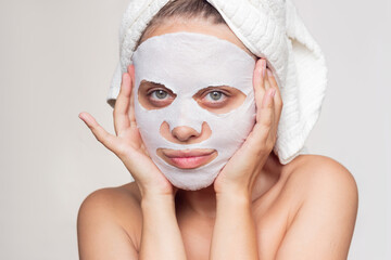 Wall Mural - A young beautiful caucasian smiling woman with a white towel on her head after a shower puts a fabric cosmetic moisturizing mask on her face on a whire background. Skin care, cosmetology