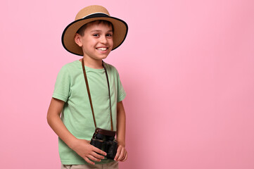 Handsome and cheerful schoolboy with a retro vintage camera around the neck smiles looking at the camera while posing over pink background with copy space