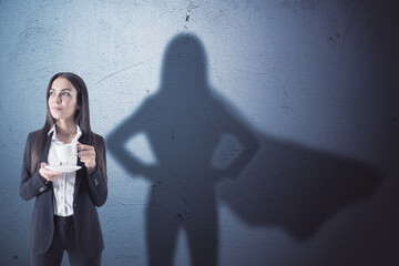 Wall Mural - Attractive young european businesswoman with coffee cup standing on concrete wall background with superhero cape shadow. Success and leadership concept.