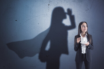 Wall Mural - Attractive young caucasian businesswoman with coffee cup standing on concrete wall background with superhero cape shadow. Success and leadership concept.
