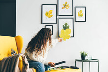 Young Woman With Long Loose Curly Hair Puts Dried Leaves Applique On Frame Glass And Covers With Sheet Of Paper Against Decorated Wall At Home Low Angle Shot