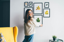 Skilled Woman Designer With Long Loose Curly Hair Holds Wooden Frame With Dried Leaf Applique And Hangs On Decorated Wall In New Apartment Backside View.