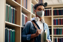 Portrait Of Black Male Student Wearing Protective Face Mask In Library And Looking At Camera.