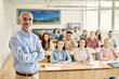 © Drazen - Happy mature teacher stands with arms crossed in classroom full of university students.