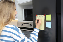 Young woman writing To do list on refrigerator door in kitchen