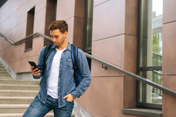 Wall Mural - Medium shot of handsome delivery man with large thermo backpack holding phone, looking at screen, using navigation app. Courier male getting order standing on stairs of office building.