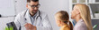 © LIGHTFIELD STUDIOS - Doctor looking at child while pointing at laptop near smiling parent, banner