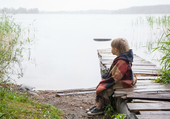 the boy is sitting on the pier by the lake, wrapped in a blanket, and looks into the distance. Fog. The concept of travel. copy space.