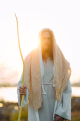 Wall Mural - Jesus Christ with wooden staff stands in white robe near river at sunset.
