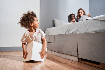 Wall Mural - Girl sitting at the floor at the bedroom and looking at her parents while playing