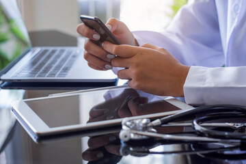 Doctor hand holding and using mobile smart phone and work on laptop computer with digital tablet and medical stethoscope on the table at office in hospital. Medical technology, telehealth concept.