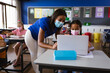 © WavebreakMediaMicro - African american female teacher wearing face mask teaching a girl to use laptop at elementary school