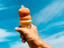 Colorful Orange Ice Cream Cone Held By Mans Hand Against Bright Blue Sky And White Clouds.