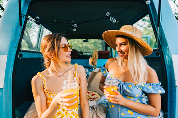 Wall Mural - Two attractive cheerful women drinking lemonade near van, enjoying summer vibes in road trip