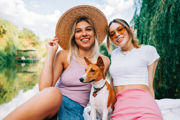 Wall Mural - Portrait of two cheerful women friends resting outdoor on lake pier with dog.