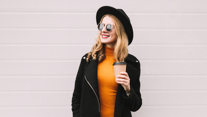 Wall Mural - Portrait of stylish happy smiling young woman with cup of coffee and looking away wearing a black coat, round hat on gray wall background