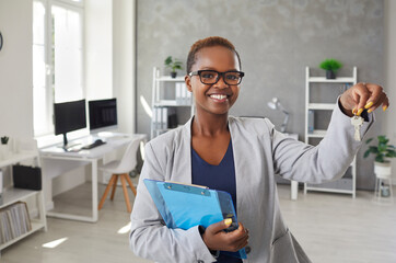 Portrait of happy real estate agent or mortgage broker in suit and glasses showing key to new house. Beautiful short haired young black woman standing in office, holding clipboard and keys and smiling