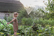 A little girl with pigtails in a sundress stands under the spray of a watering pad in the middle of a vegetable garden in summer