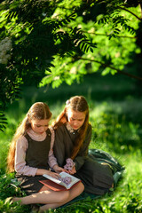 Wall Mural - Two schoolgirls in school uniforms are reading a book while sitting on the green grass in the park. Do homework