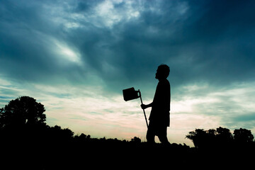 Wall Mural - a silhouette of a person holding a long tool, standing in a field at sunset. There are trees and clouds in the background.