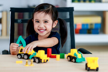 Little Cute Pigtails Hairstyle Preschooler Kindergarten Happy Girl Sit On Chair Smiling And Playing Backhoe Loader Truck Brick Toy On Table In Living Room At Home In Front Of Bookshelf