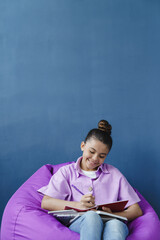 Wall Mural - Positive teenage girl sitting in a comfortable pouf, studying with a smile. Little girl on a blue background