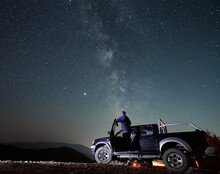 Rear View Of Young Man Who Enjoying Magic Milky Way And Many Stars In Night Sky Looking Out Of The Cabin Black SUV Car. Night Riding On Stony Mountain Roads By Big Car.