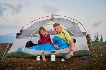 Wall Mural - Young man in sleeping bag in tent preparing morning coffee for his beloved, waking up after cool night in the mountain fresh air. Tourist breakfast outdoors. Concept of camping and relationships.