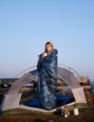 girl standing in open tent, wrapped around in tourist sleeping bag for hiking, drinking warm tea and